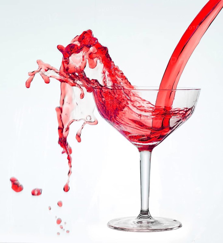A powerful flood of red liquid pours into a cocktail glass and spills over, studio shot against a white background