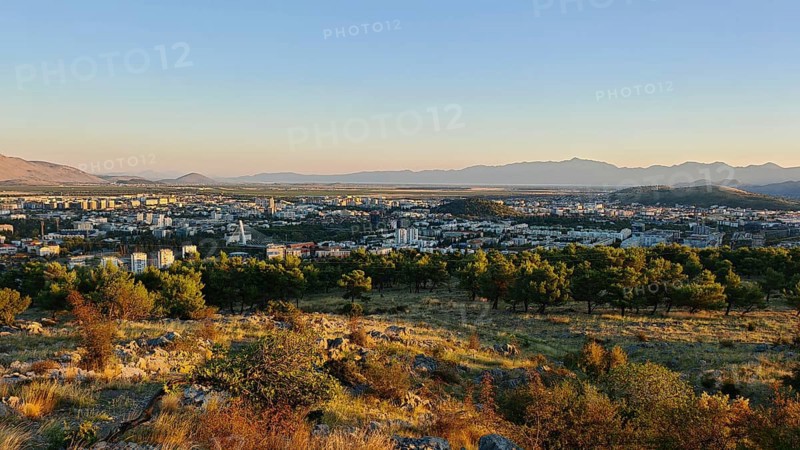 A city surrounded by nature with trees in the foreground and mountains in the background, illuminated by a warm sunset, looking out from a hill on the outskirts of Podgorica, Montenegro