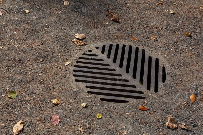 Close-up of heavy cast iron metal storm drain cover on compacted gravel path with fallen leaves in autumn, Quebec, Canada