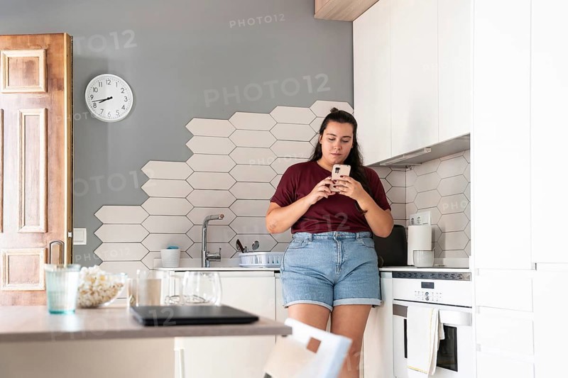 A curvy woman in a casual outfit stands in a modern kitchen, intently using her smartphone. The stylish kitchen features hexagonal tiles and a clock