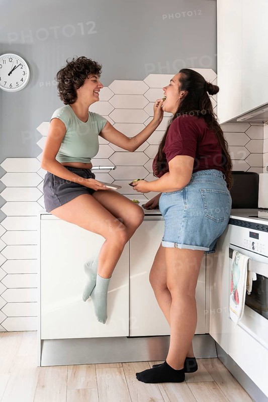 A happy lesbian couple shares a playful moment in a modern kitchen. One partner sits on the counter, feeding the other with a smile, showcasing a warm, intimate connection