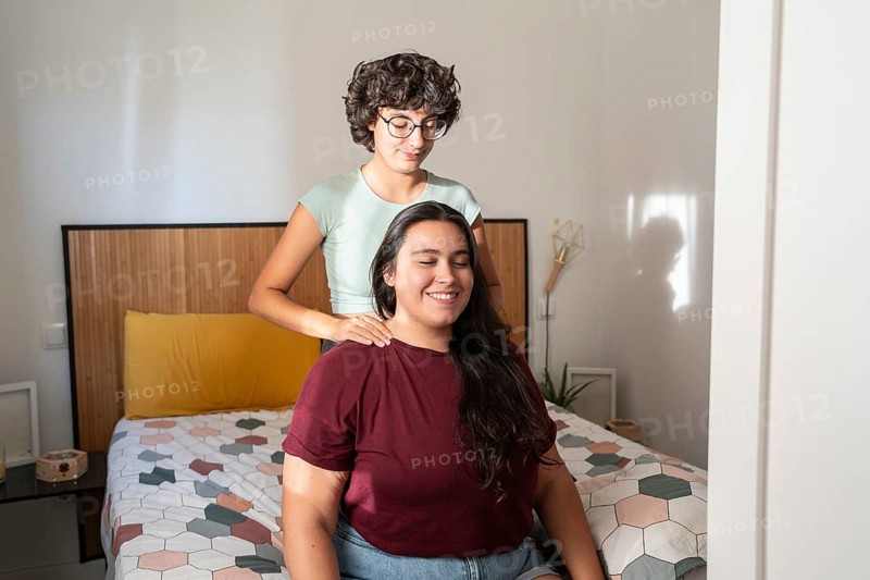 A loving lesbian couple sharing a peaceful moment in a cozy bedroom, with one partner gently massaging the other's shoulders, creating a serene and intimate atmosphere