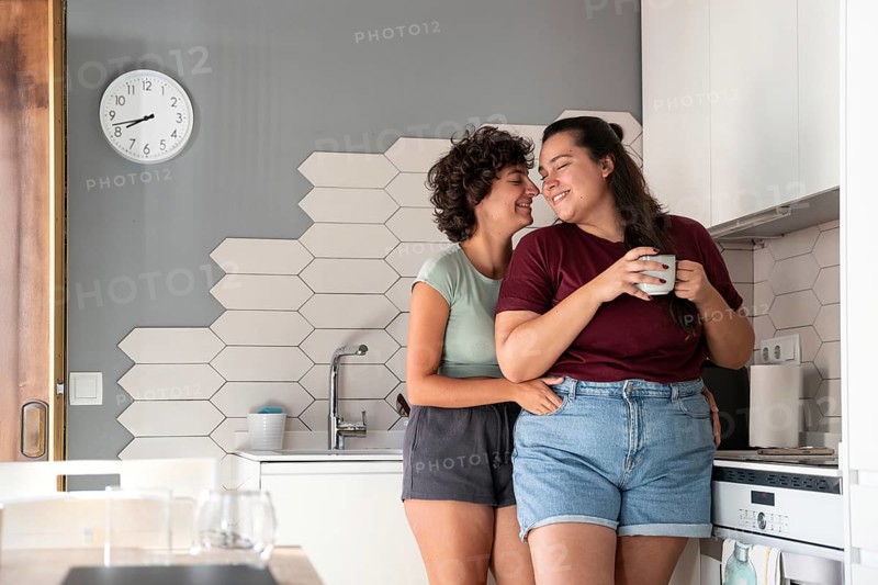 A happy lesbian couple shares a playful moment in a modern kitchen. One partner sits on the counter, feeding the other with a smile, showcasing a warm, intimate connection