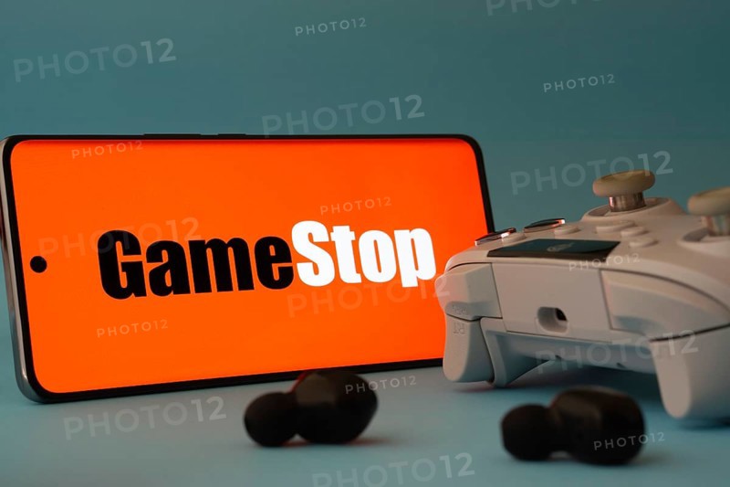 Dhaka, Bangladesh- 15 July 2025: A smartphone with a GameStop icon next to a controller and earbuds on a blue surface