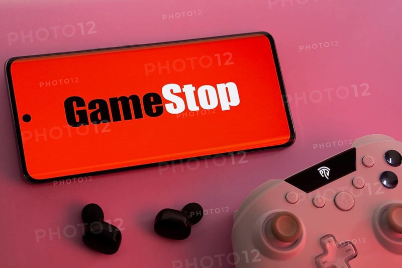 Dhaka, Bangladesh- 15 July 2025: Smartphone with GameStop logo, game controller, and earbuds on a pink background