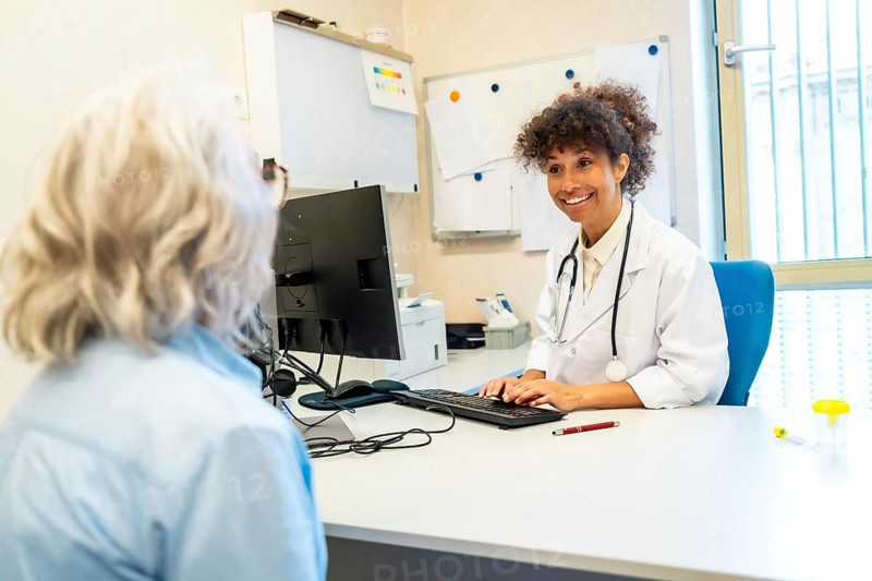 Smiling female doctor wearing lab coat and stethoscope typing on computer keyboard while listening to senior female patient in hospital room