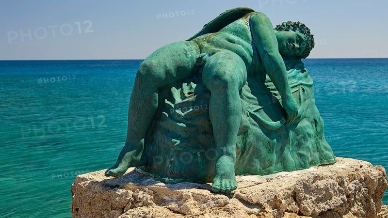Bronze sculpture, sleeping Eros, close-up of an artistic statue on a rock by the blue water, harbour area, Rhodes Town, Rhodes, Dodecanese, Greek Islands, Greece