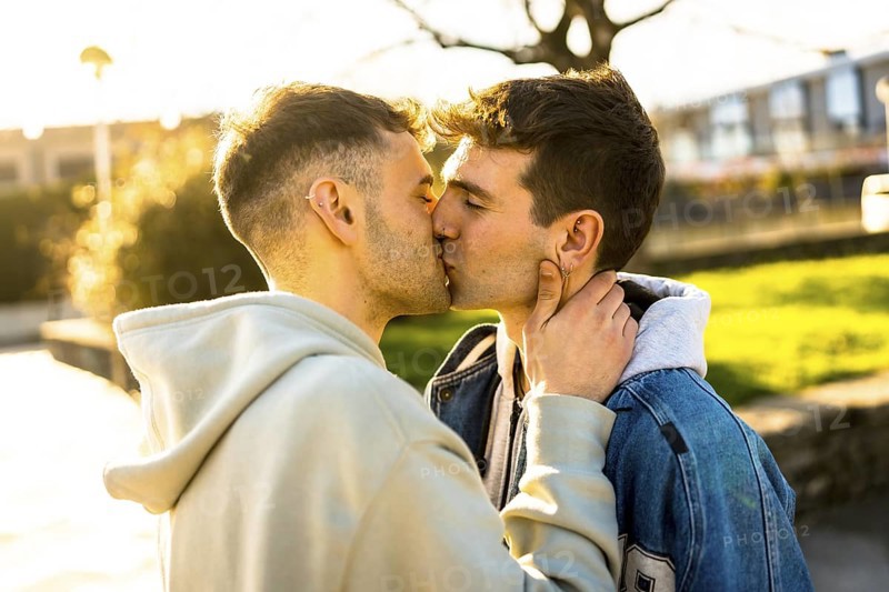 Romantic gay couple sharing a tender kiss in a park, enveloped in the warm glow of a breathtaking sunset, celebrating their love