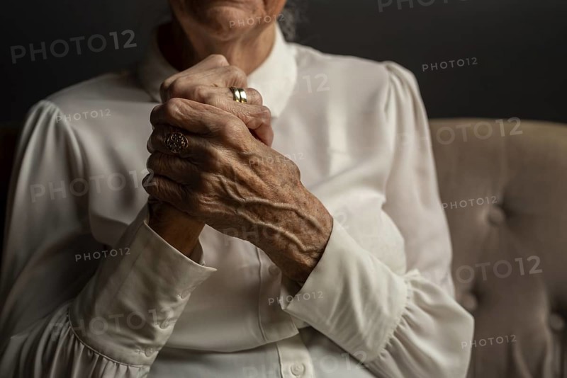 90 year old woman in white blouse holding folded hands, conveying thoughtfulness in a dim light, old age, symbolic image, Germany