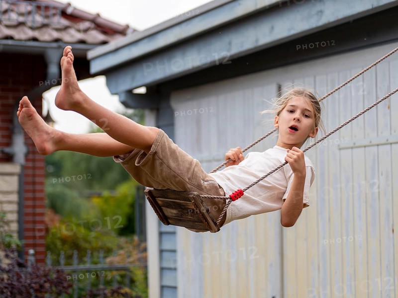 Boy swinging while sitting on children's swing at shed and garden gate, Germany