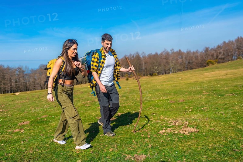 Young couple walking hand in hand across a vibrant green meadow, carrying backpacks and using a hiking stick under a sunny sky