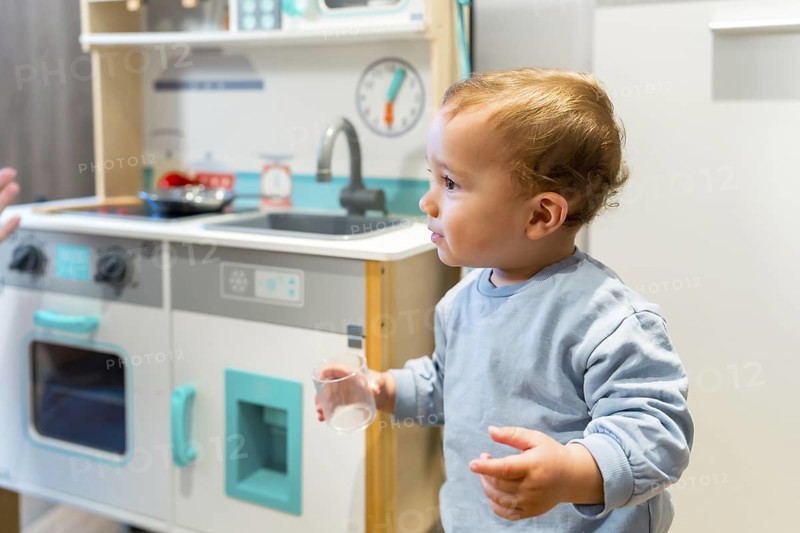 Curious toddler holding a glass while playing with toy kitchen set, exploring and learning through imaginative play