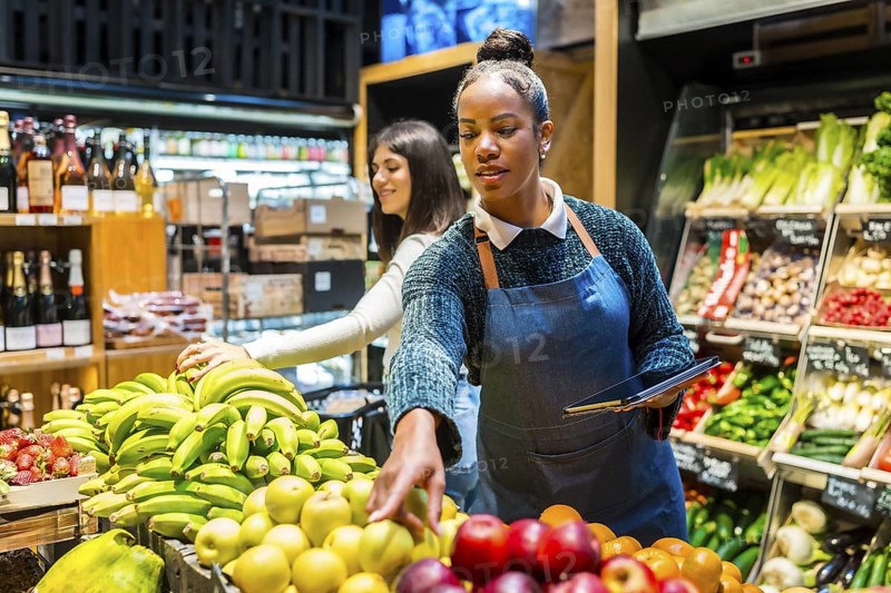 Supermarket employee using digital tablet while arranging fresh fruit on display, customer choosing bananas in background