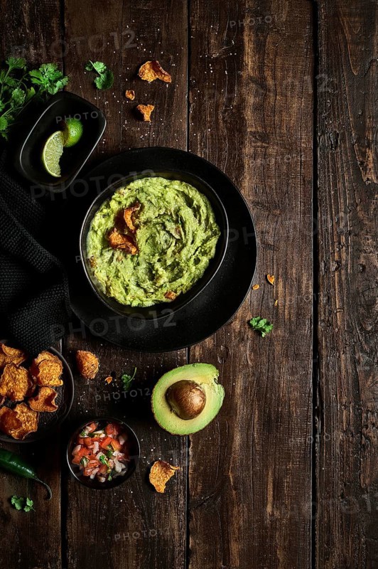 Top view of bowl of fresh guacamole placed near avocado and chips on weathered lumber tabletop