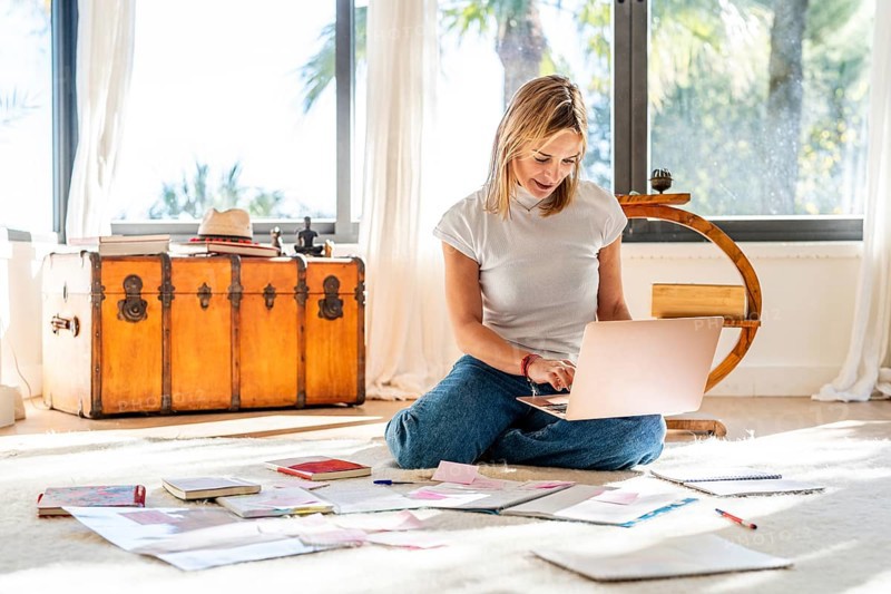 A woman sits on a bright home floor, absorbed in her laptop, with papers scattered around her. Sunlight streams through large windows