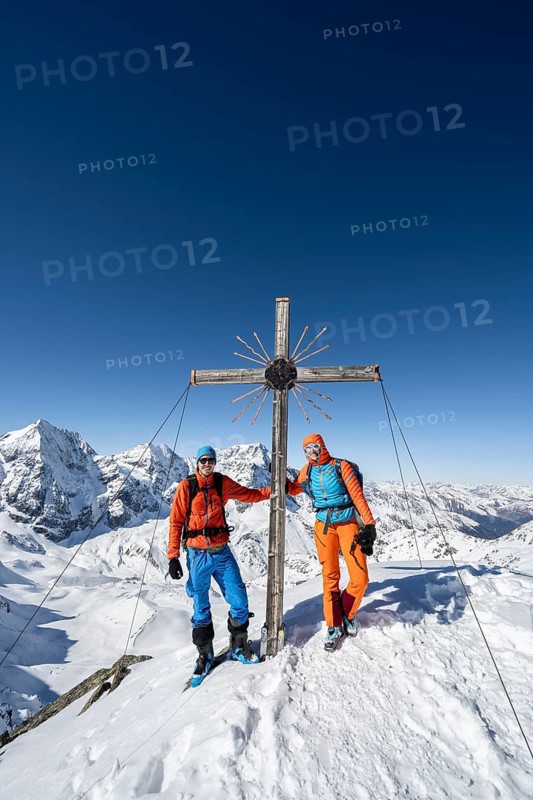Two ski tourers at the summit cross on the summit of the Madritschspitze, mountain panorama with snow-covered mountain landscape in winter, view of mountain peaks Königsspitze, Monte Zebru and Ortler, Ortler Alps, Vinschgau Valley, Italy