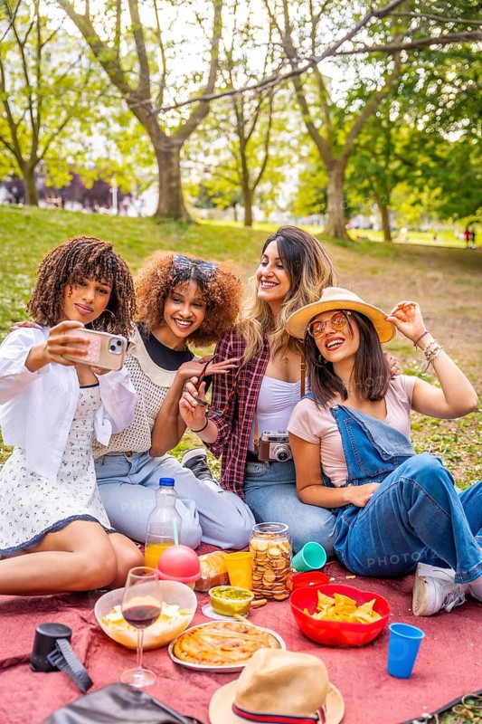 Four cheerful young women enjoying a sunny picnic in the park, happily taking a selfie with their smartphone while surrounded by nature