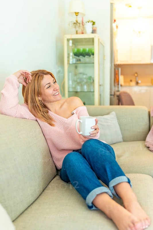 Vertical photo of a blonde mature adult happy relaxed woman drinking tea lying on the sofa