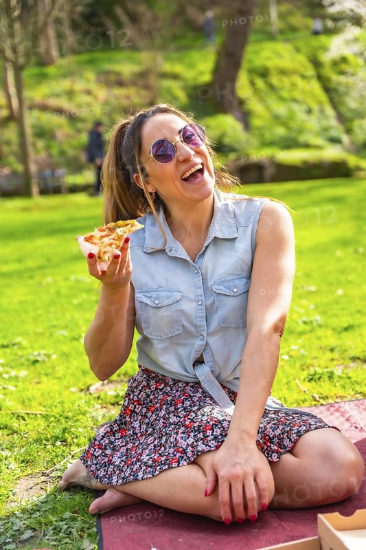 Cheerful young woman laughing while eating pizza in a vibrant green park, enjoying a sunny spring day