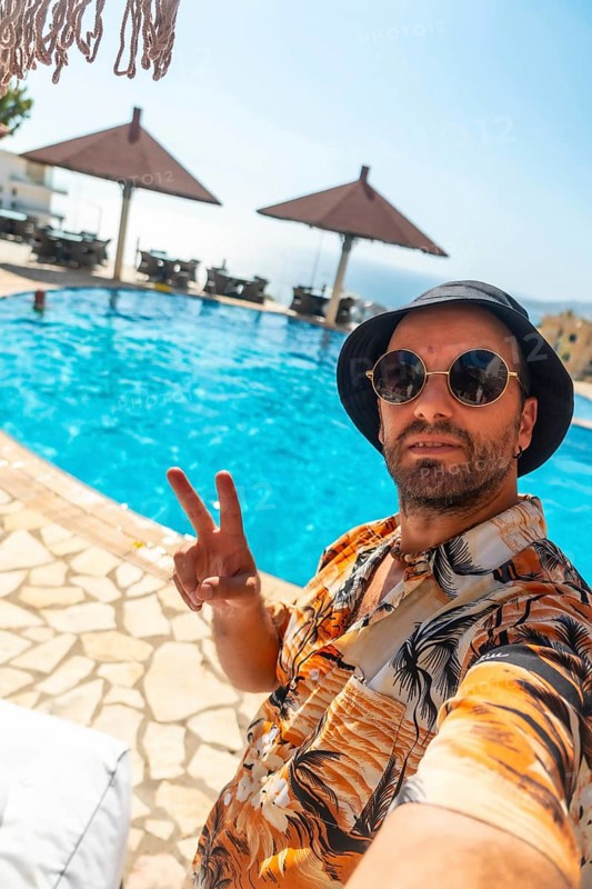Tourist man in the hotel pool in summer taking a selfie enjoying the vacation, making the victory symbol