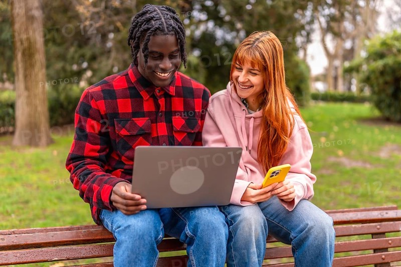 Two happy young students are using a laptop and a smartphone while sitting on a bench in a park, enjoying online learning and connectivity