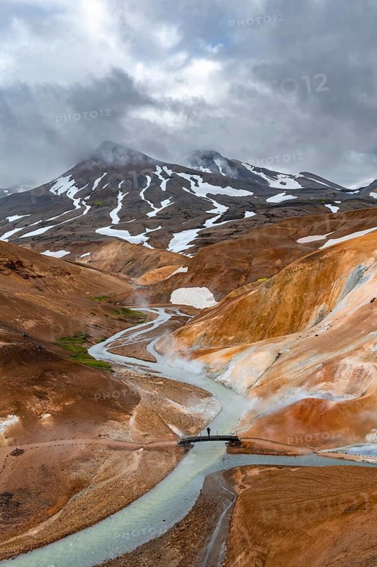 Tourist on bridge over steaming stream between colourful rhyolite mountains with snowfields, Hveradalir geothermal area, Kerlingarfjöll, Icelandic highlands, Iceland