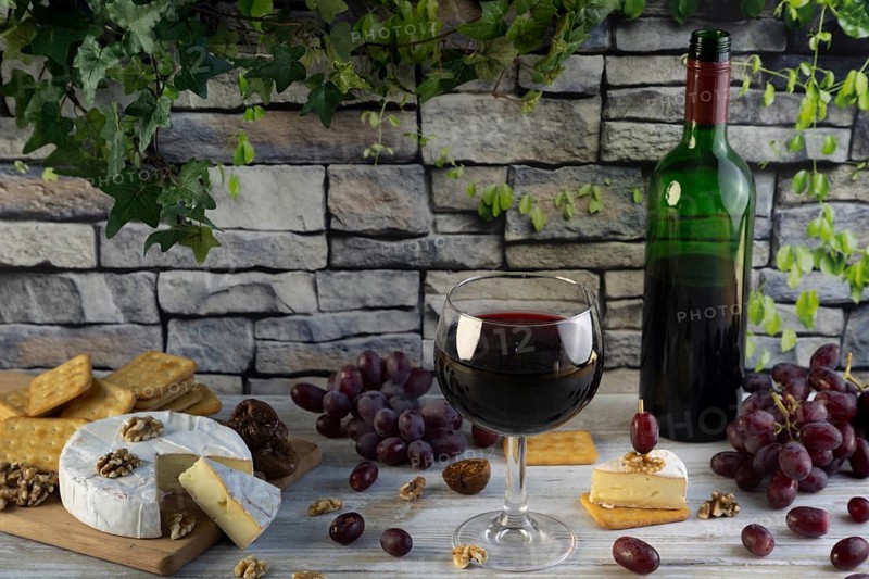 Wine glass and bottle of red wine surrounded by cheese, grapes, nuts and crackers in front of a rustic stone wall