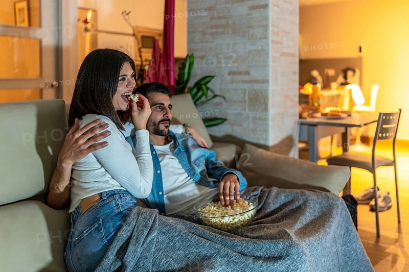 Young couple enjoying a movie night at home, eating popcorn from a bowl and relaxing on the sofa under a blanket
