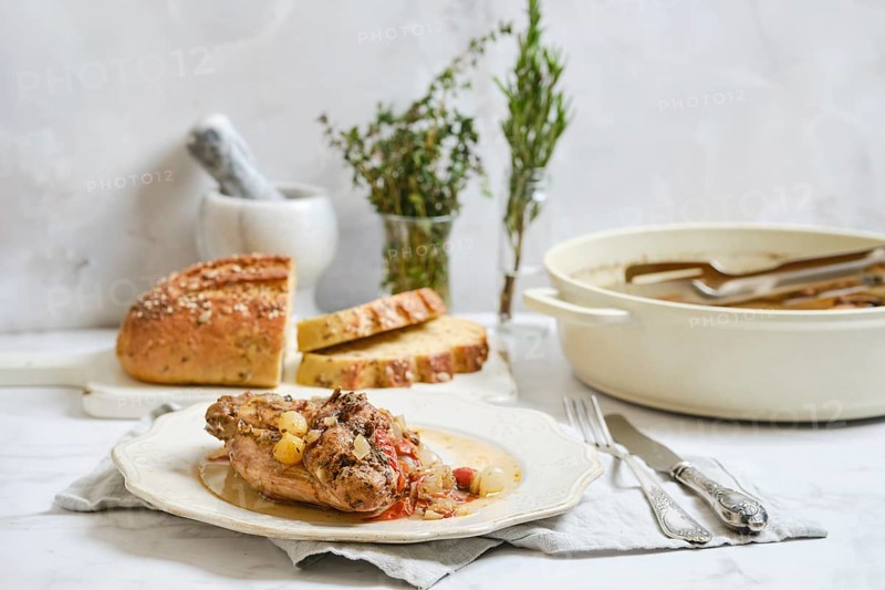Cooked rabbit stew is served on a plate with herbs on top. Fresh bread is placed next to the dish on a simple cloth. A pot is shown nearby with more stew, creating a homey kitchen scene
