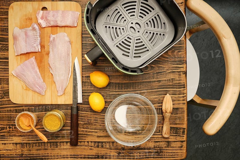 Fish fillets are arranged on a cutting board next to a bowl and spices. Fresh lemons rest nearby, with a sharp knife on the wooden table, indicating meal preparation in a home kitchen