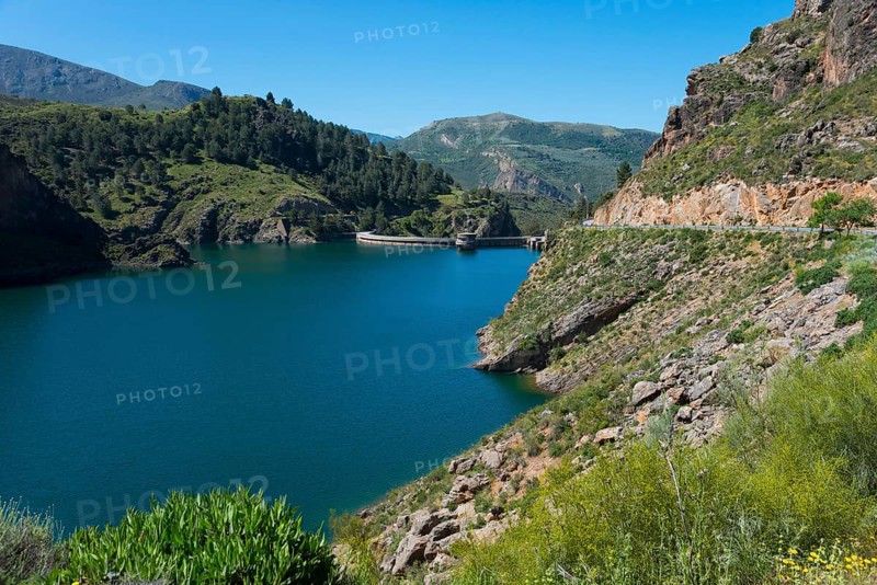 A tranquil lake in a mountainous landscape with green hills and rocks, under a clear blue sky, reservoir, Embalse de Quéntar, Quentar, Granada province, Andalusia, Spain