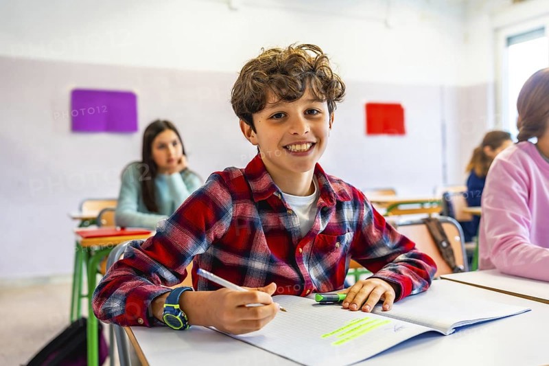 Happy elementary school student smiling and writing in notebook during class, with classmates in background