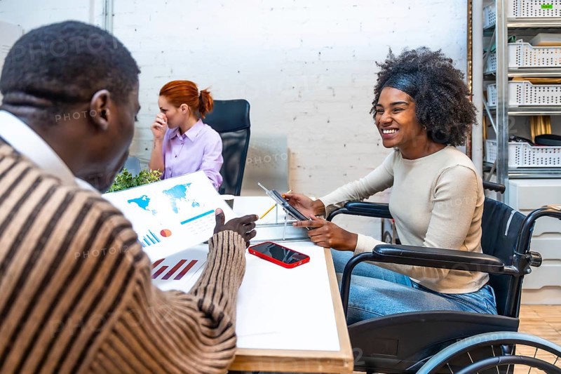 Cheerful and smiling african woman with disability sitting on wheelchair and colleague discussing graphs in a coworking