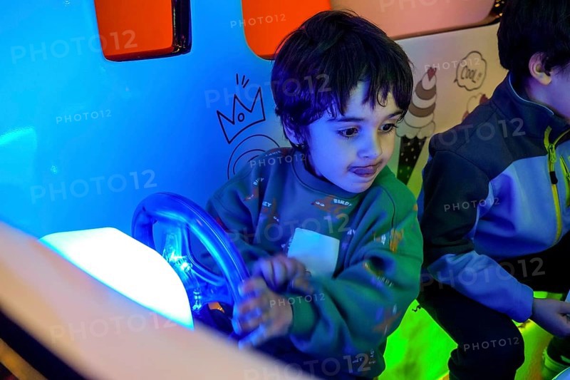 Young boy playing with toy car steering wheel, enjoying entertainment in colorful amusement park illuminated with neon lights