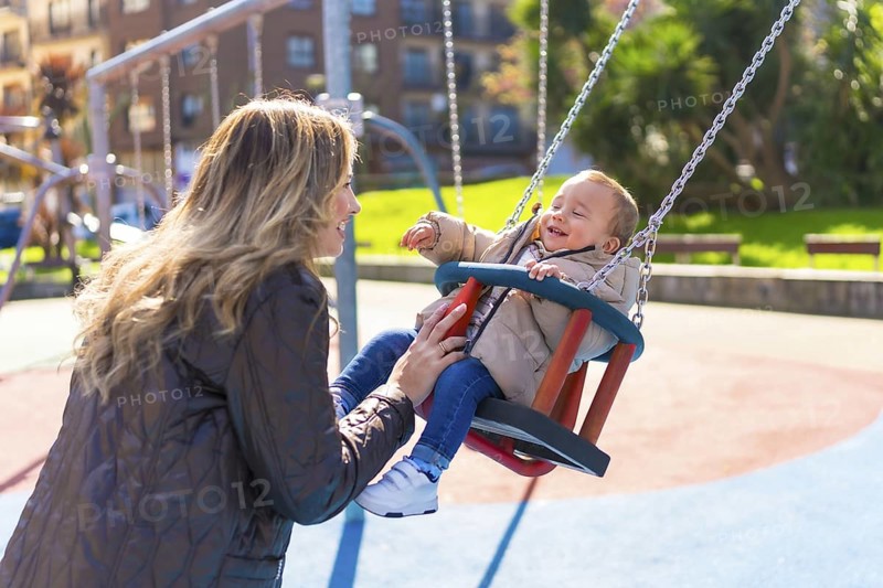 Happy mother pushing her laughing baby son on a swing, enjoying a sunny autumn day at the playground, creating joyful family memories