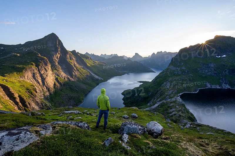 Mountaineer looking over mountain landscape with pointed mountain peaks and fjord Forsfjorden with village Vindstad, sun star at sunrise, Moskenesøya, Lofoten, Nordland, Norway