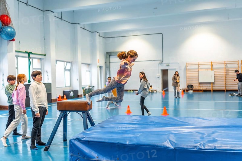 Elementary school girl practicing gymnastics, jumping over a pommel horse in the school gym watched by her classmates and teacher