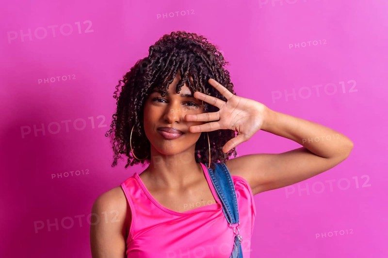 Young woman with curly hair and pink top partially covering her face with her hand on a vibrant pink background