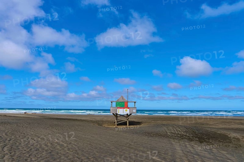 A rescue tower stands alone on a wide sandy beach under a blue sky, Bethells Beach, Te Henga, Auckland, New Zealand