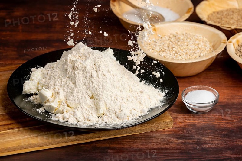 Flour is sprinkled over curd cheese, surrounded by baking ingredients on a wooden table