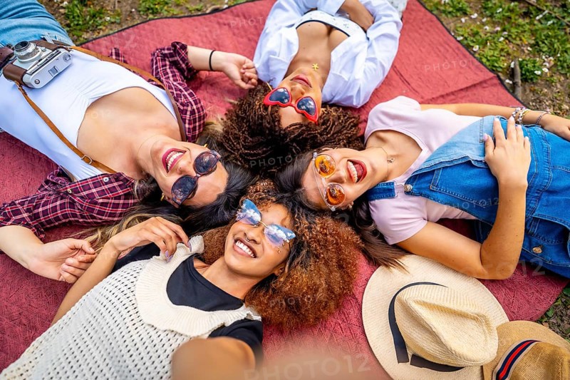 Four cheerful young women with sunglasses and hats are lying in circle on a blanket at a park, taking a selfie
