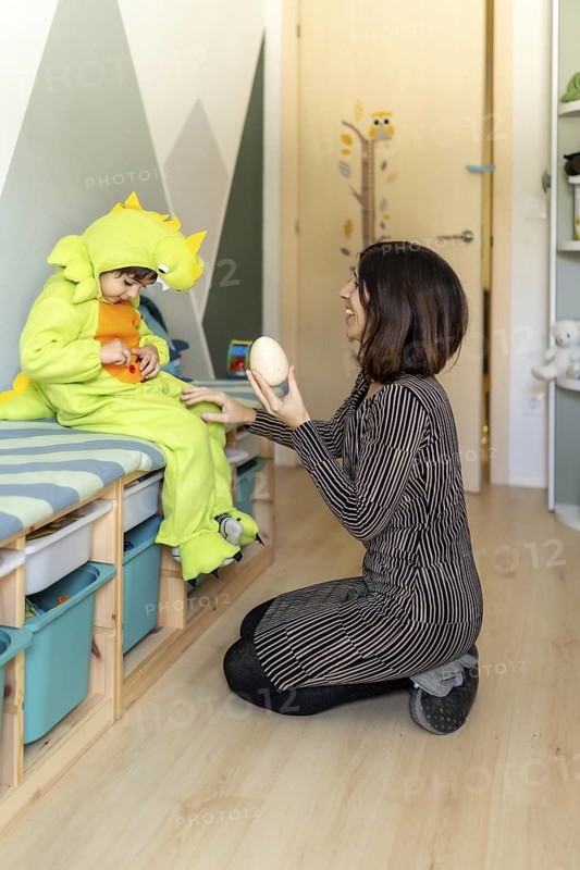 Mother kneeling and showing a dinosaur egg to her son wearing a dinosaur costume while playing in his bedroom