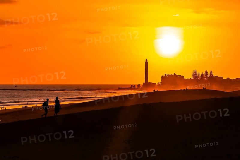 Tourists walking on the maspalomas dunes at sunset, with the maspalomas lighthouse in the background, gran canaria, canary islands, spain