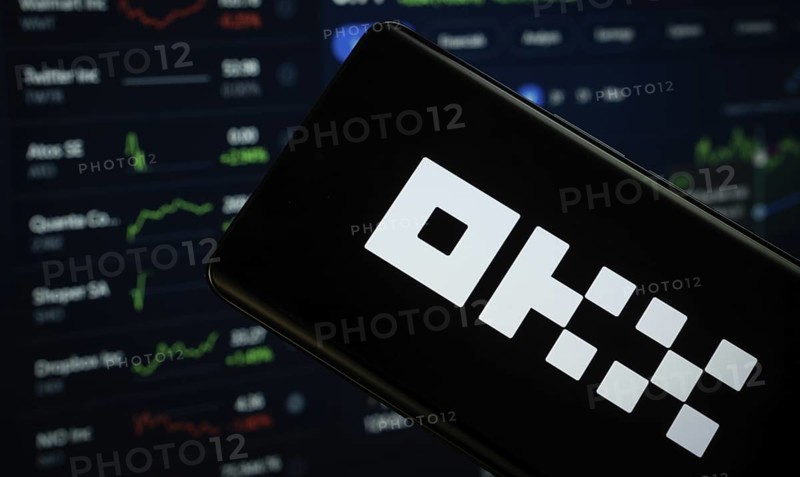 Dhaka, Bangladesh- 17 March 2025: okx logo is displayed on a smartphone screen, with a stock graph webpage visible on a computer screen in the background
