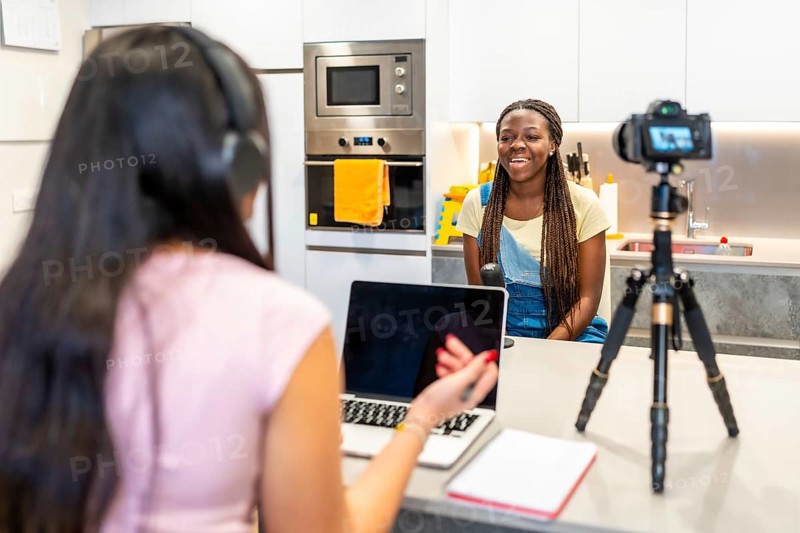 Two teenage girls creating content for their video blog, recording with a camera and using a laptop in a home kitchen