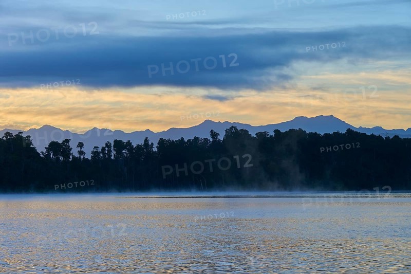 A hazy lake at sunrise with a mountainous silhouette and blue surroundings, Lake Mahinapua, Ruatapu, South Island, New Zealand