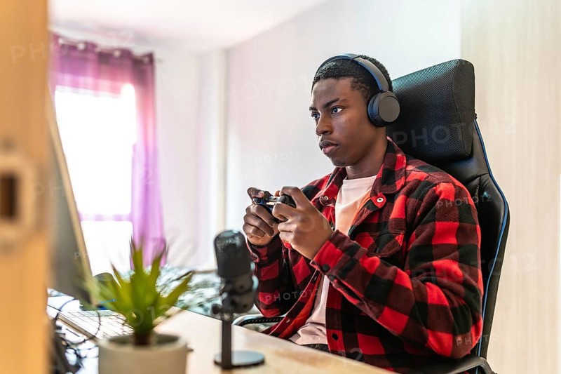 Young man playing videogames on a desktop computer using a controller, wearing headphones and using a microphone