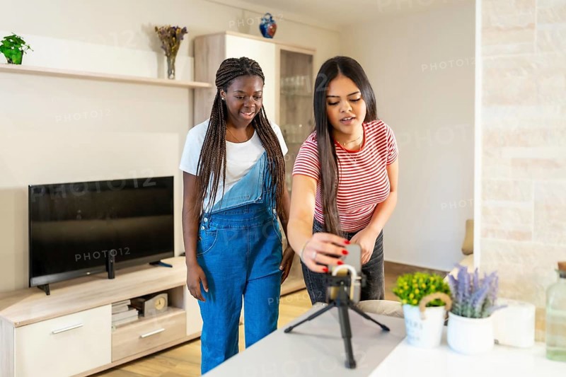 Two teenage girls preparing equipment for recording video content at home, using smartphone and microphone