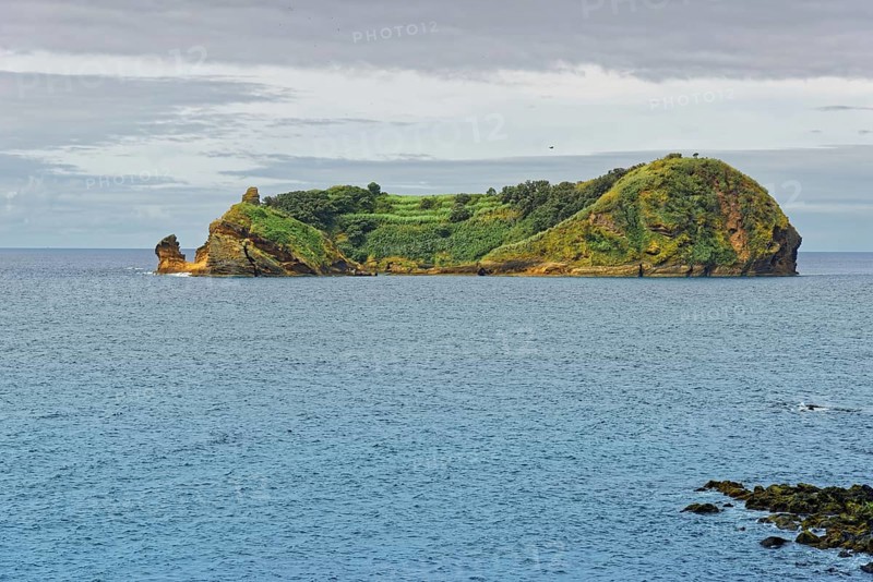 Green, rocky island and protected area Ilhéu de Vila Franca do Campoin a calm sea under a cloudy sky, Vila Franca do Campo, Sao Miguel, Azores, Portugal