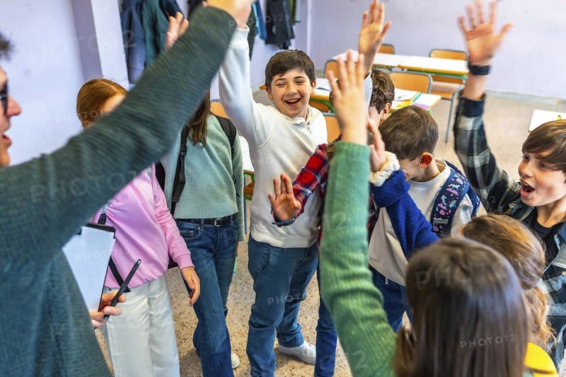 Group of cheerful elementary school students and teacher raising hands together in classroom, celebrating success and teamwork